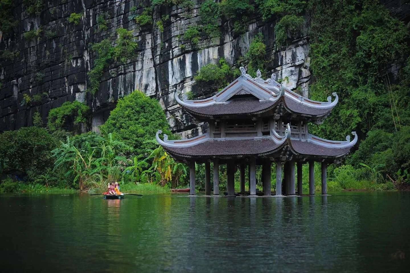 Thủy đình mái cong giữa mặt nước xanh dưới vách núi đá vôi Tràng An, Ninh Bình, có thuyền nan chở khách