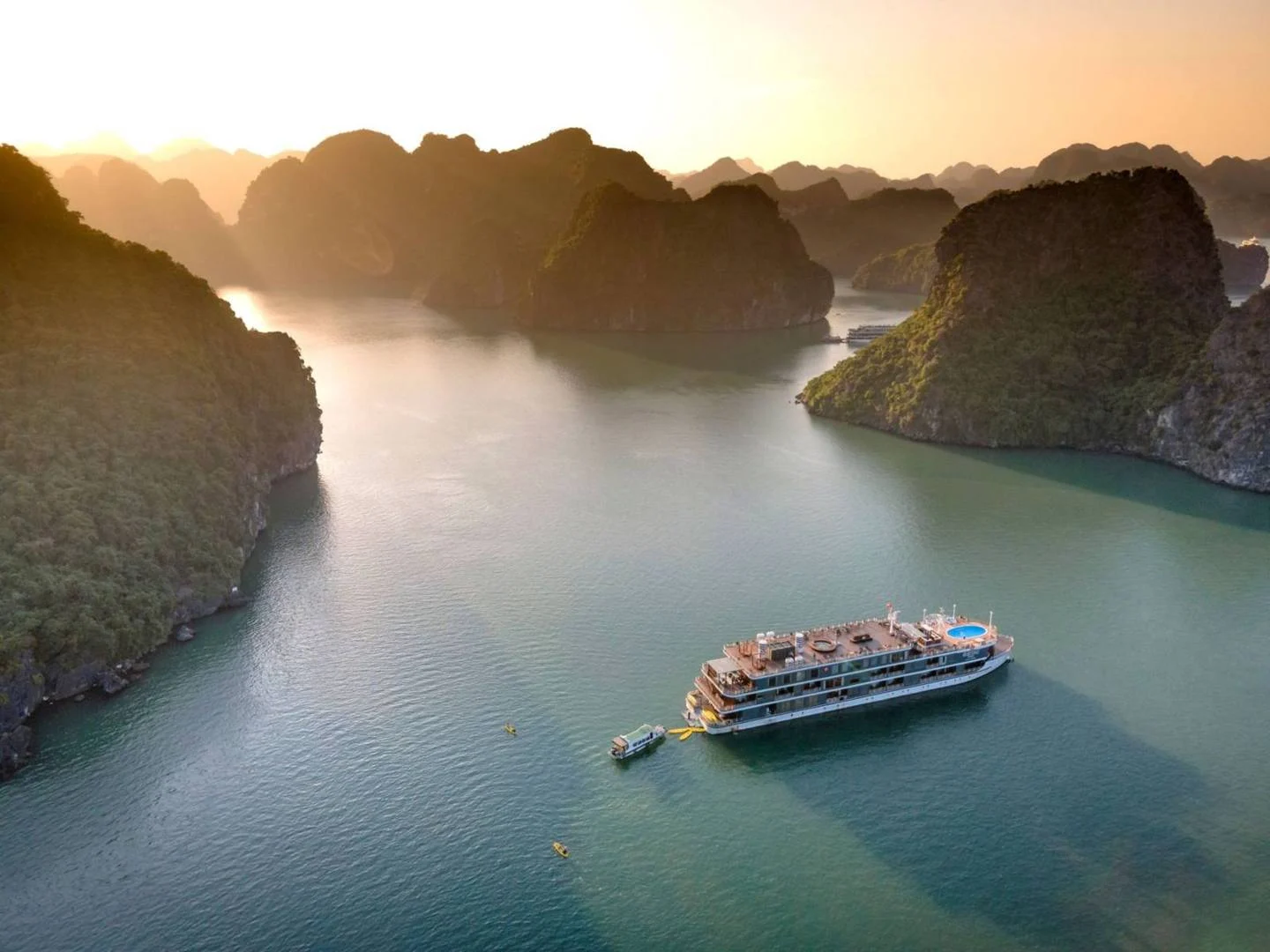 Du thuyền nhiều tầng neo giữa vịnh Hạ Long lúc hoàng hôn, xung quanh là các đảo đá vôi và thuyền kayak